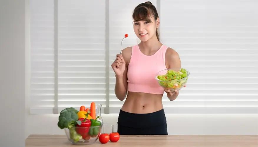 Fit woman in pink sports top eating fresh salad with vegetables on kitchen table